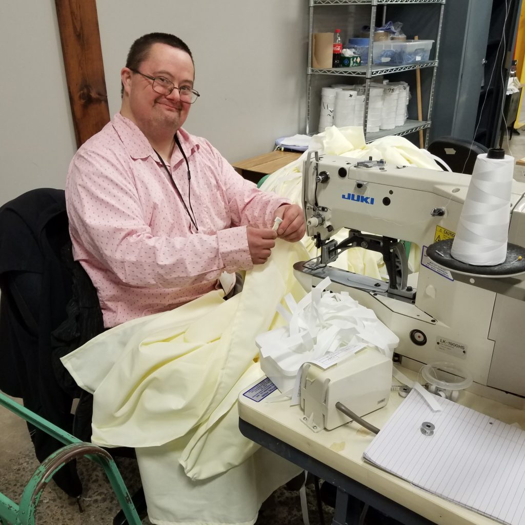 An adult man with disabilities wearing a pink button-down shirt working on sewing an isolation gown on a Jung sewing machine.