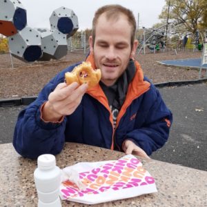 An adult man with disabilities sitting at a table in a park, holding a half-eaten donut from Dunkin' Donuts.
