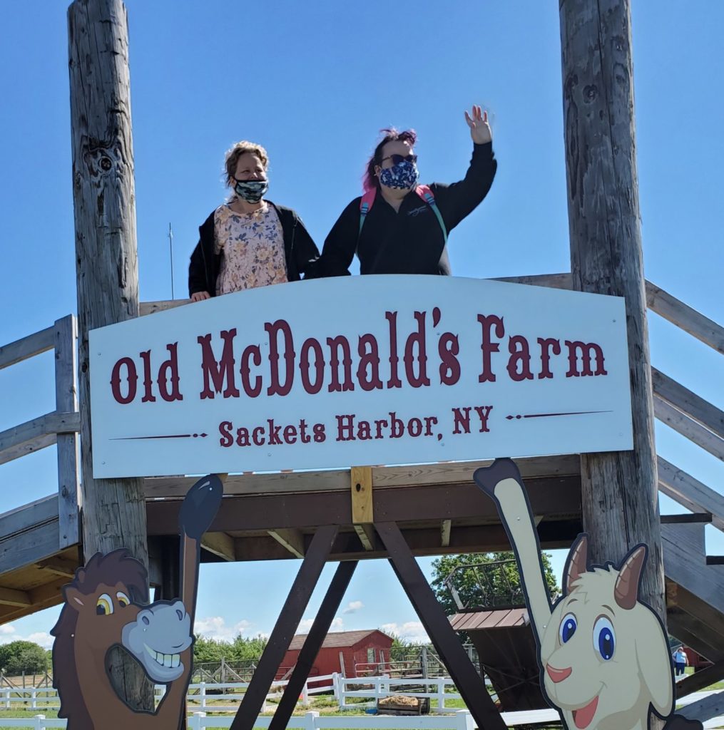 Two adult women with disabilities standing on a bridge with a sign hung from the front. The sign says, "Old McDonald's Farm, Sackets Harbor, NY." One of the two women is waving to someone off-camera to her left.