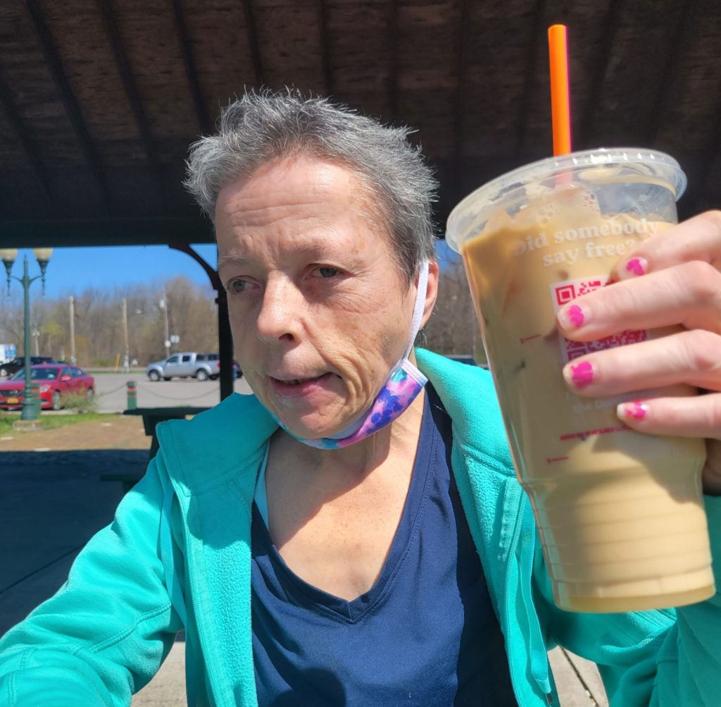 An older woman with disabilities holds up a Dunkin' iced coffee. Her face mask is pulled down below her chin and she appears to be ready to take a sip. She is sitting in a public park in a covered pavilion.
